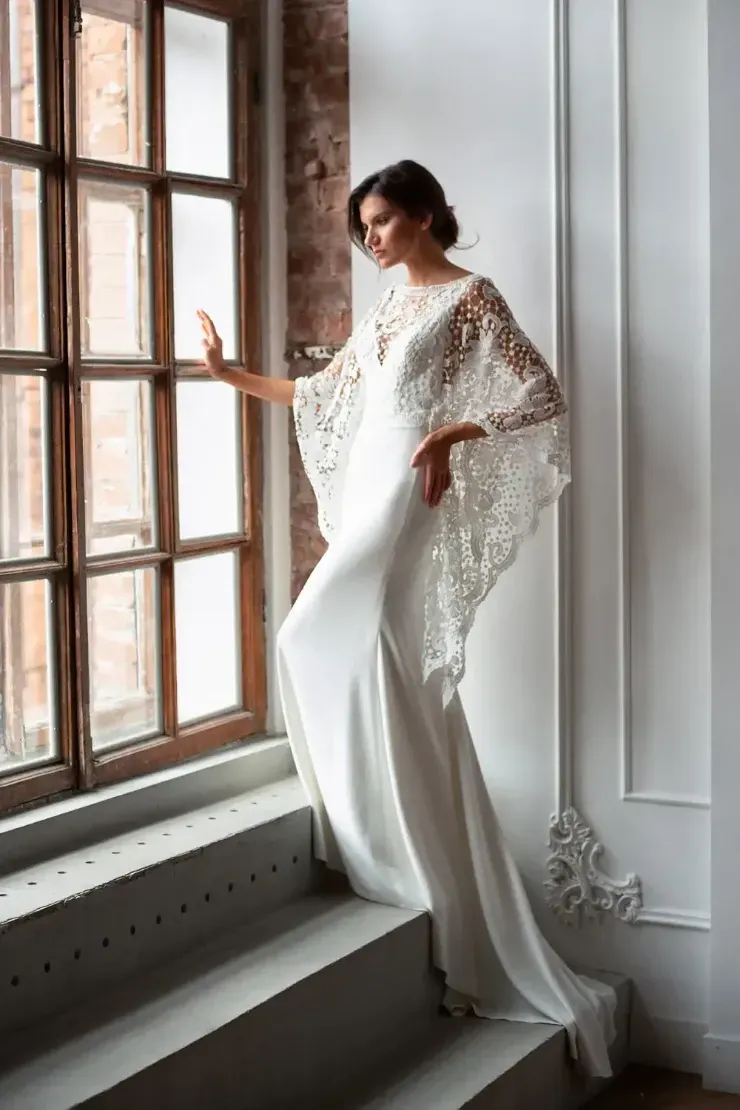 A woman in a white dress is standing next to a window.