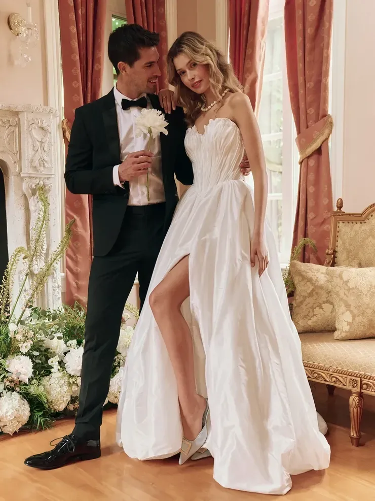 A bride and groom are posing for a picture in a room . the bride is wearing a high slit wedding dress.