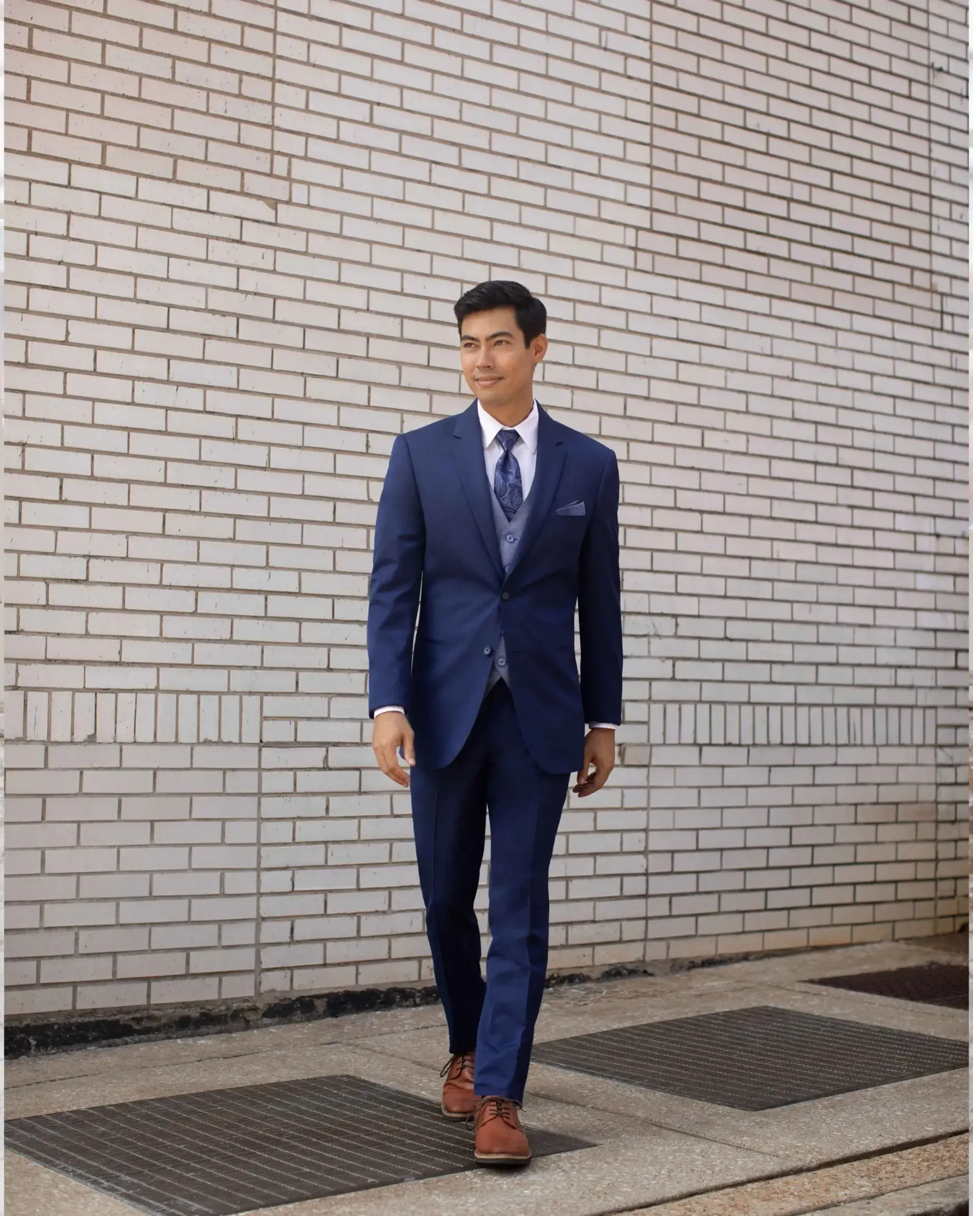 A man in a blue suit and tie is walking in front of a white brick wall