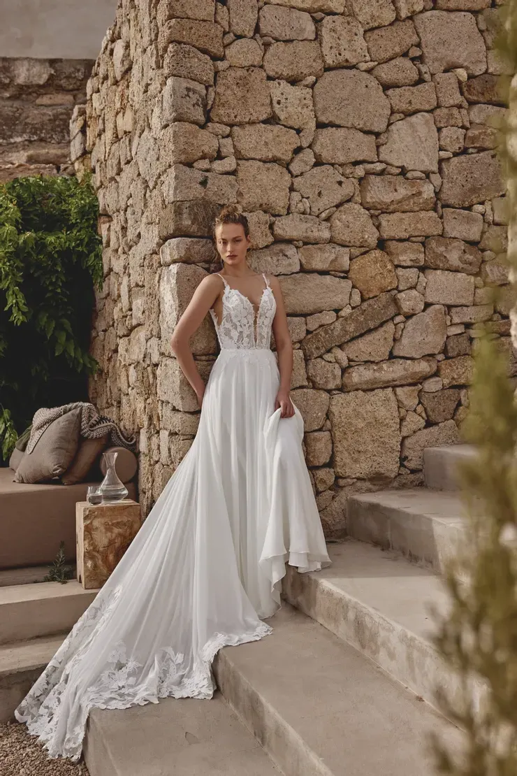 A woman in a white wedding dress is standing on stairs in front of a stone wall.
