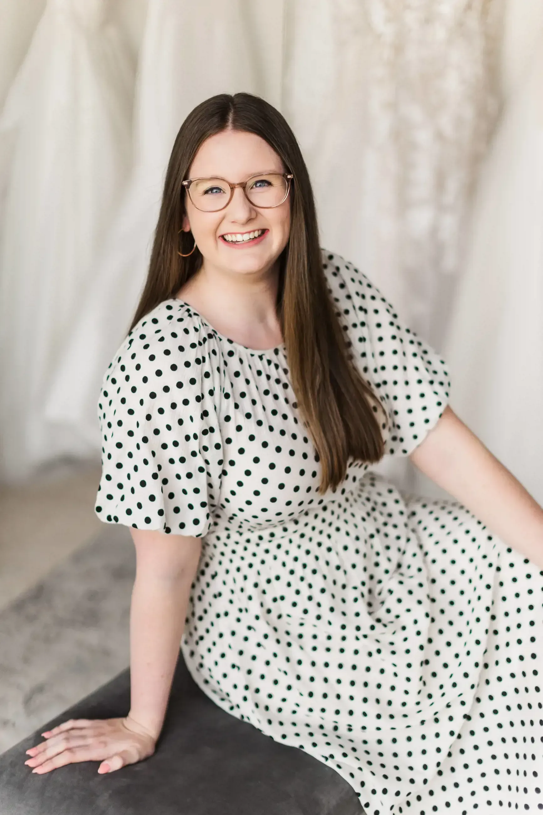 A woman in a polka dot dress and glasses is sitting on a bench.