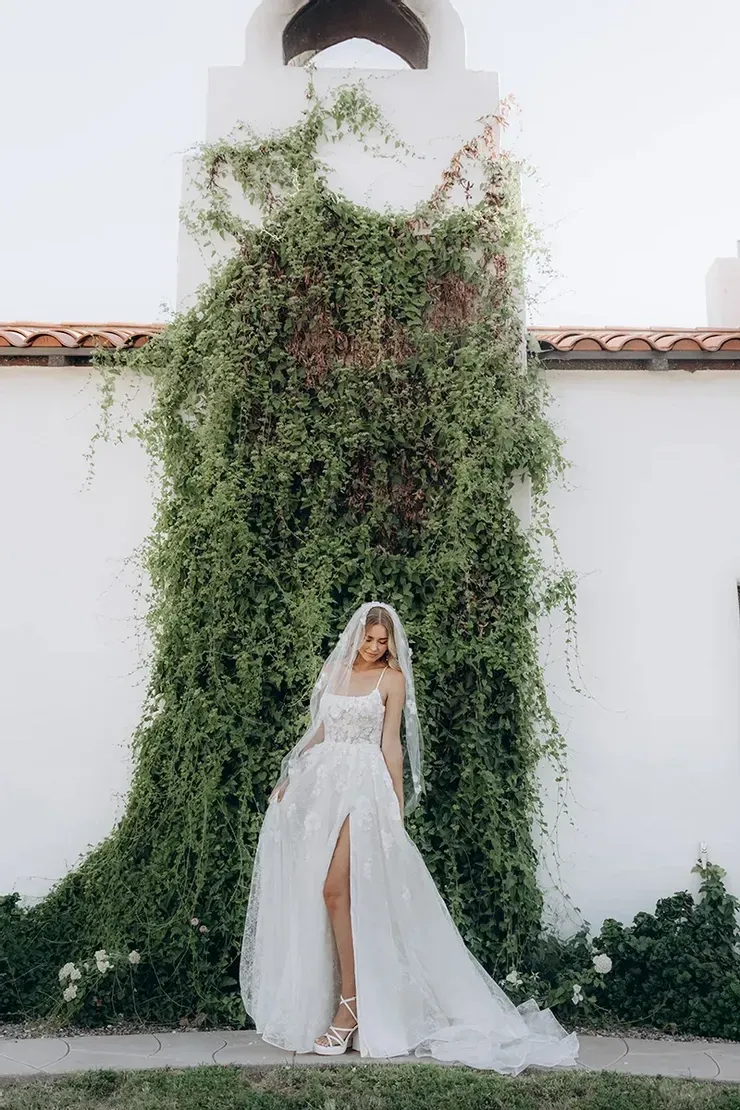 A bride in a white dress and veil is standing in front of a white building.