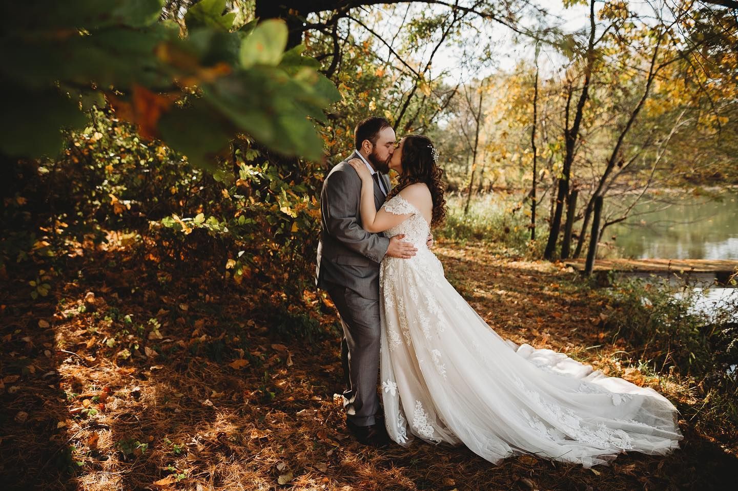 A bride and groom are kissing in the woods near a lake.