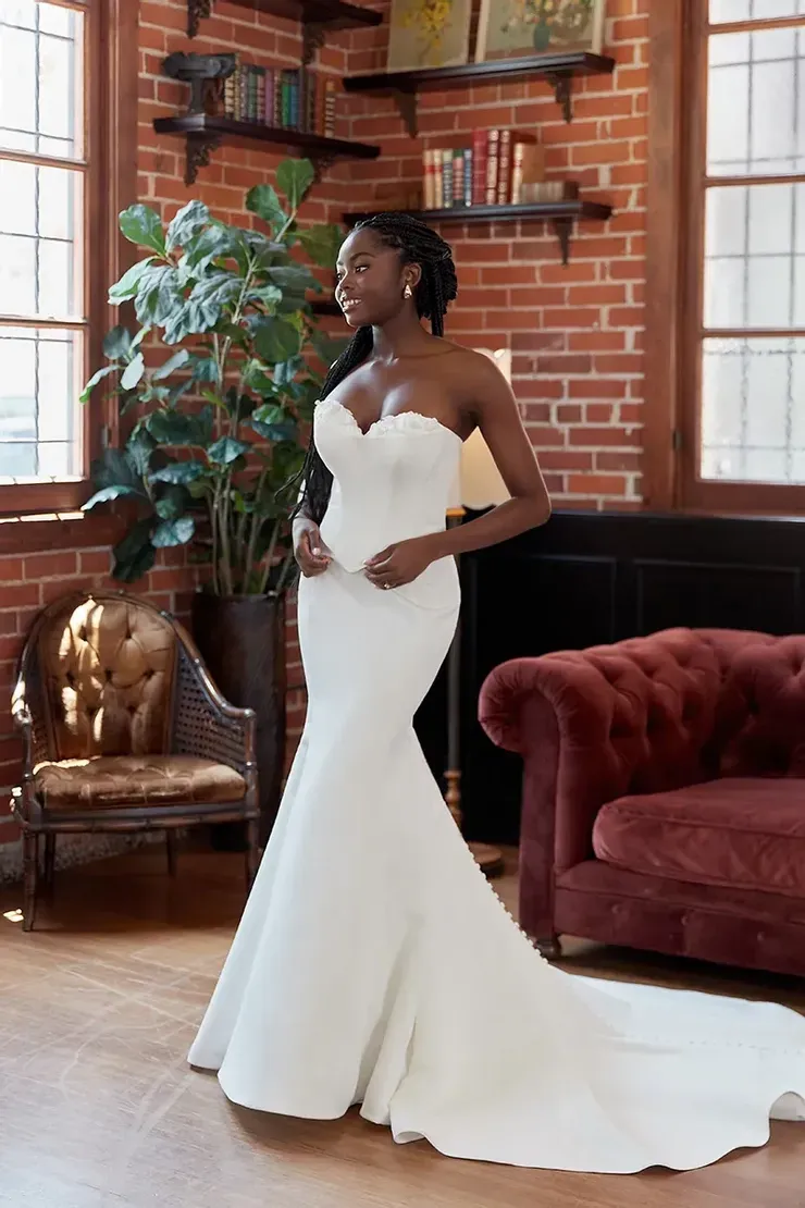 A woman in a white wedding dress is standing in a living room next to a red couch.