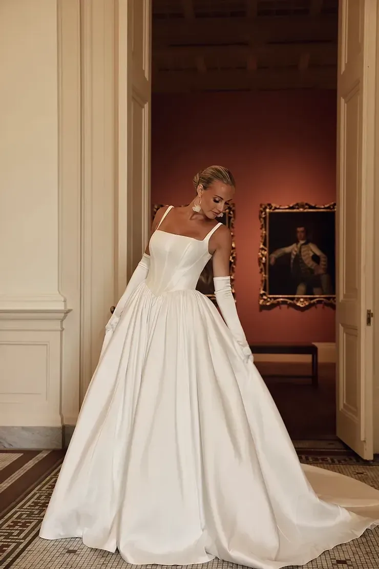 A woman in a white wedding dress and gloves is standing in a room.