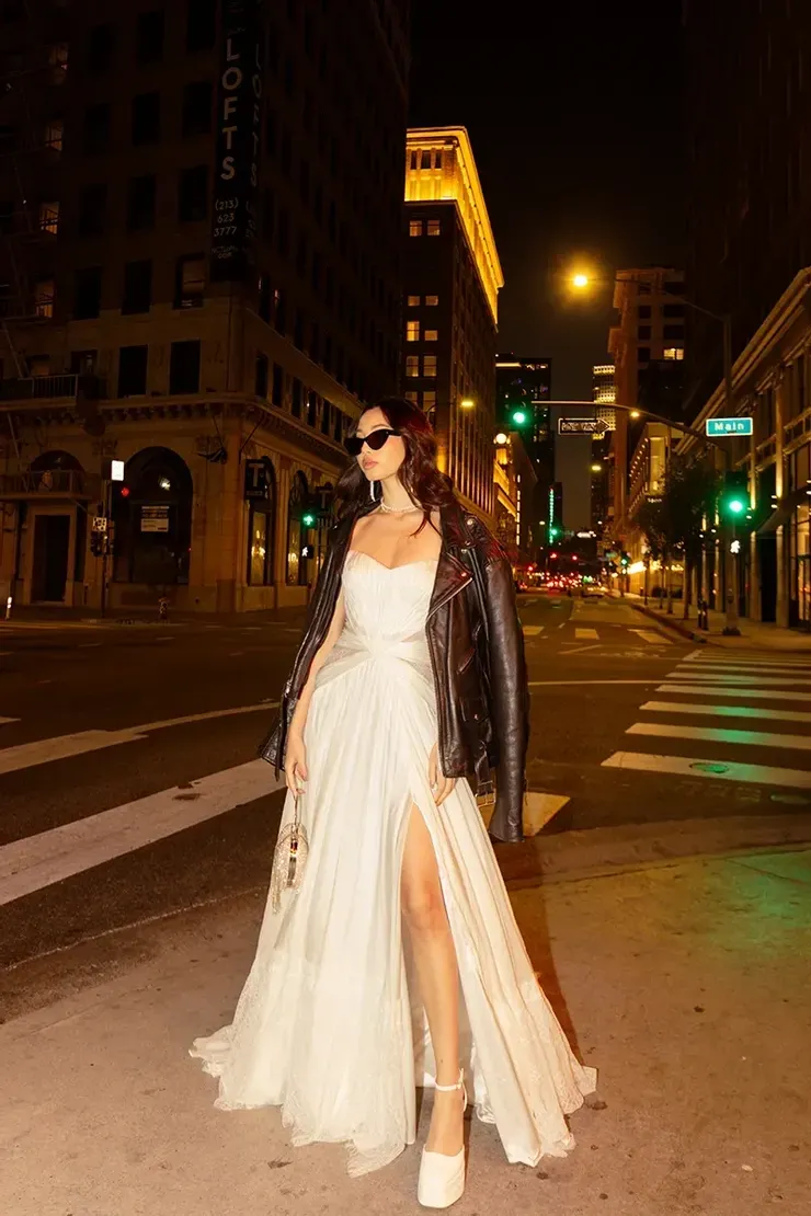 A woman in a white dress and a leather jacket is standing on a city street at night.