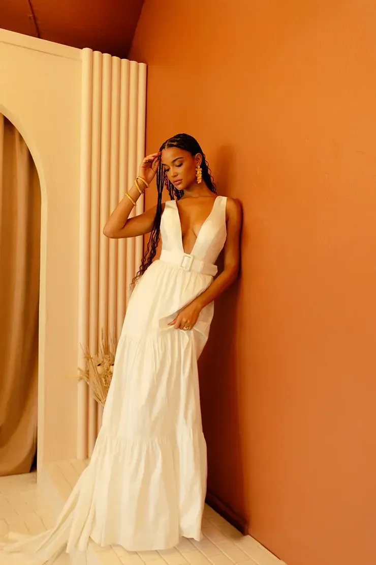 A woman in a white dress is leaning against a wall.