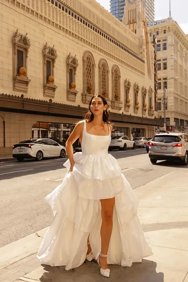 A woman in a high low wedding dress is standing on the sidewalk in front of a building.