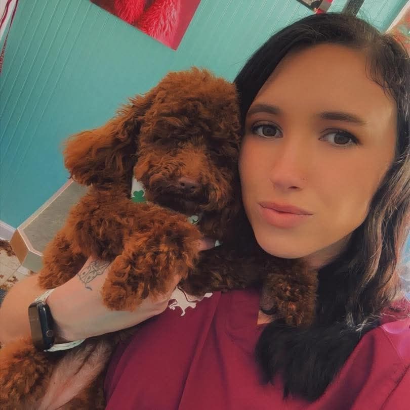 Woman holding a small curly brown dog, both close to the camera indoors.