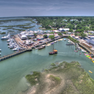 Aerial view of a coastal marina with boats, docks, waterfront buildings, and green wetlands nearby