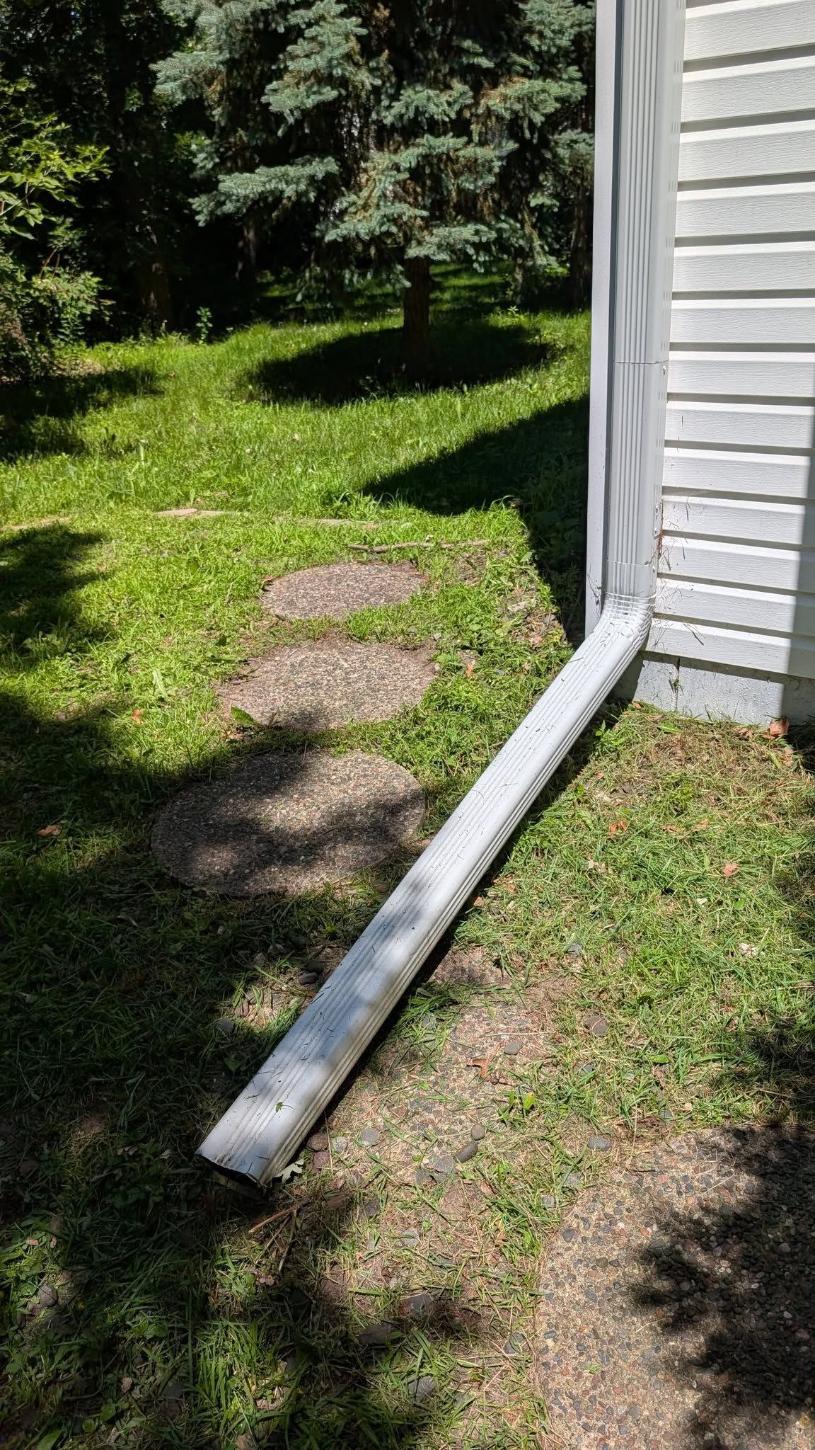 White guttering directs water from a building's corner onto a grassy area with circular stepping stones.