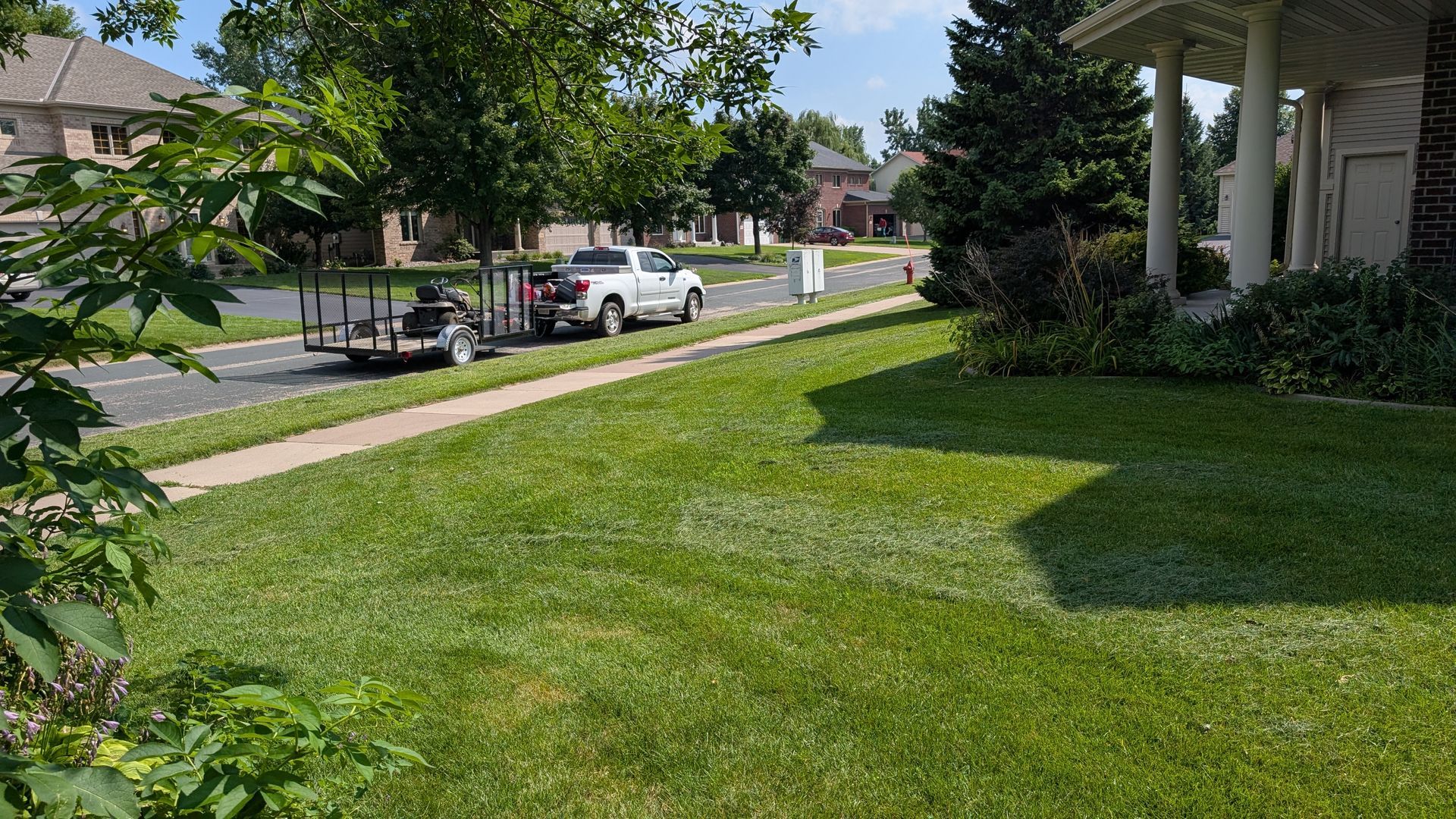 Lawn care truck and trailer on a street with a house. Green grass in foreground, trees, blue sky.