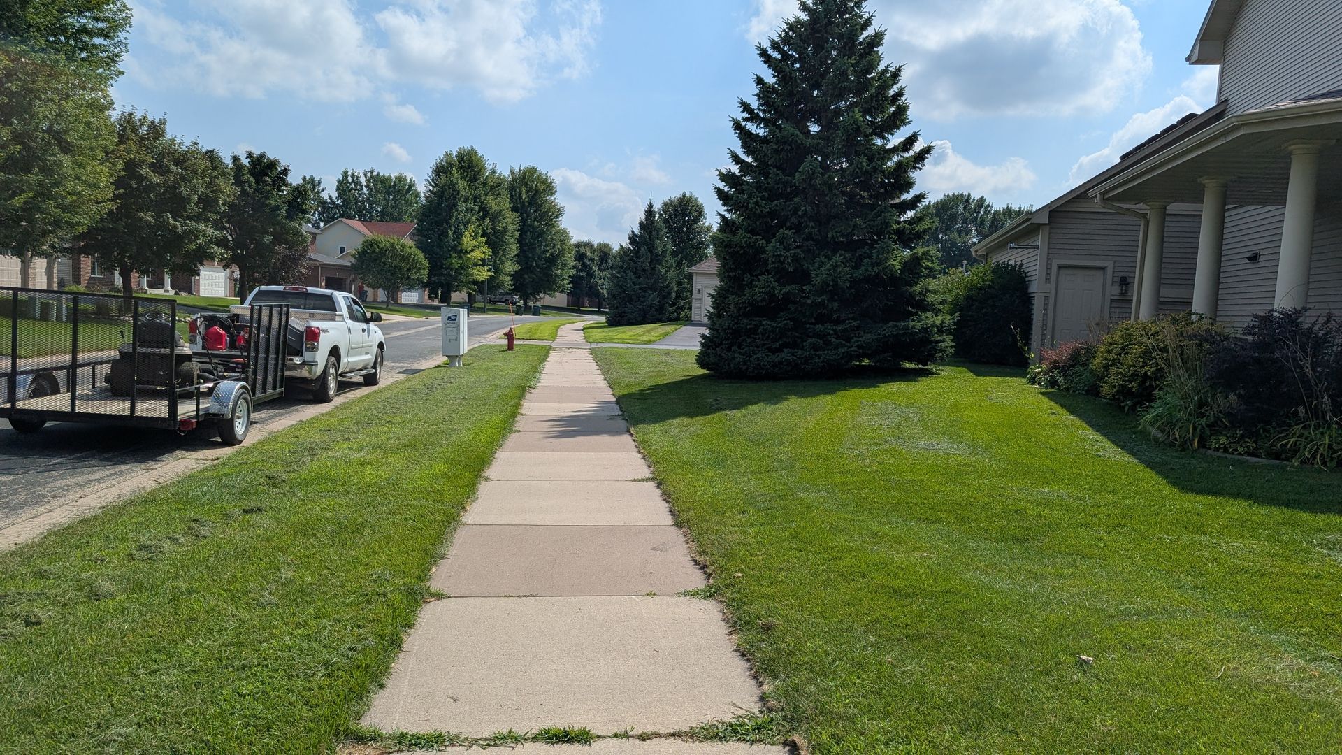 Sidewalk in a residential area, flanked by green lawns and trees, under a sunny sky.