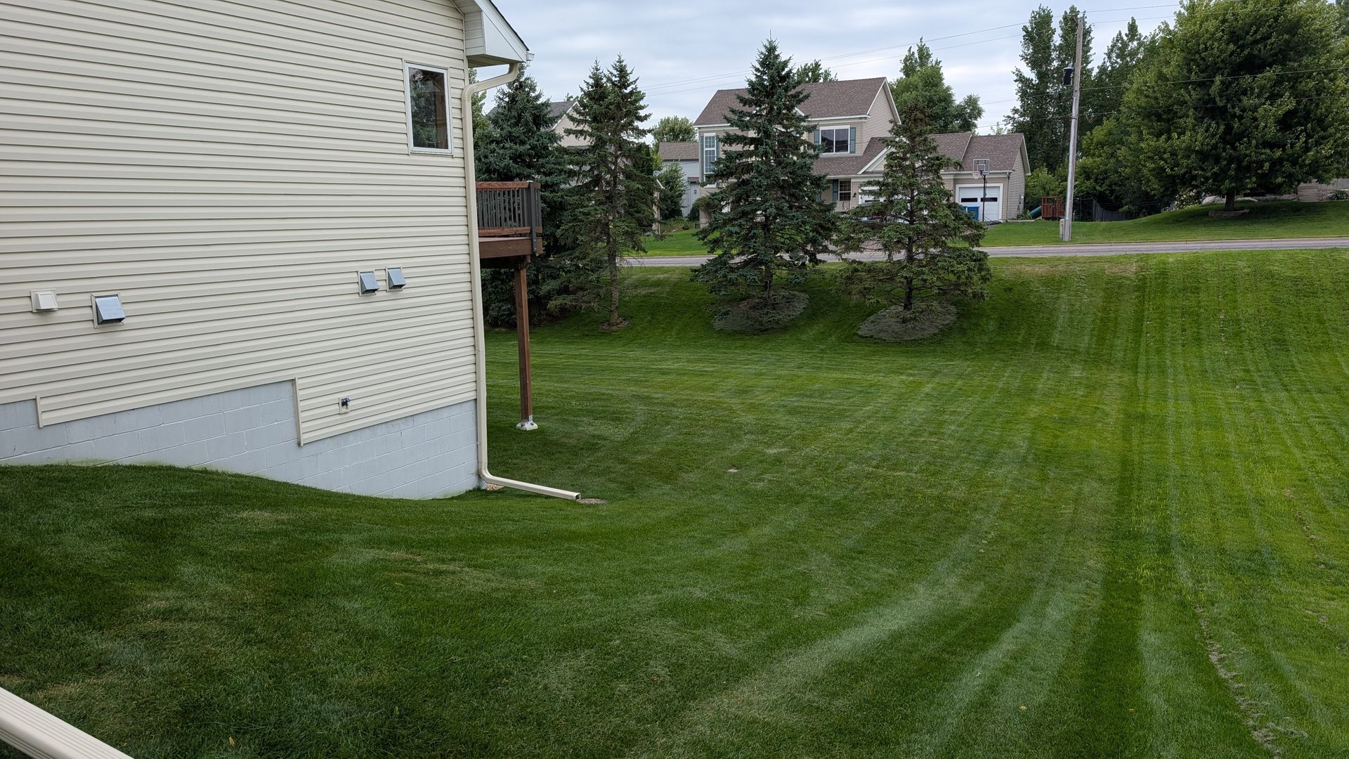 Lawn next to a beige house, green grass, a few trees, and houses in the distance under a cloudy sky.