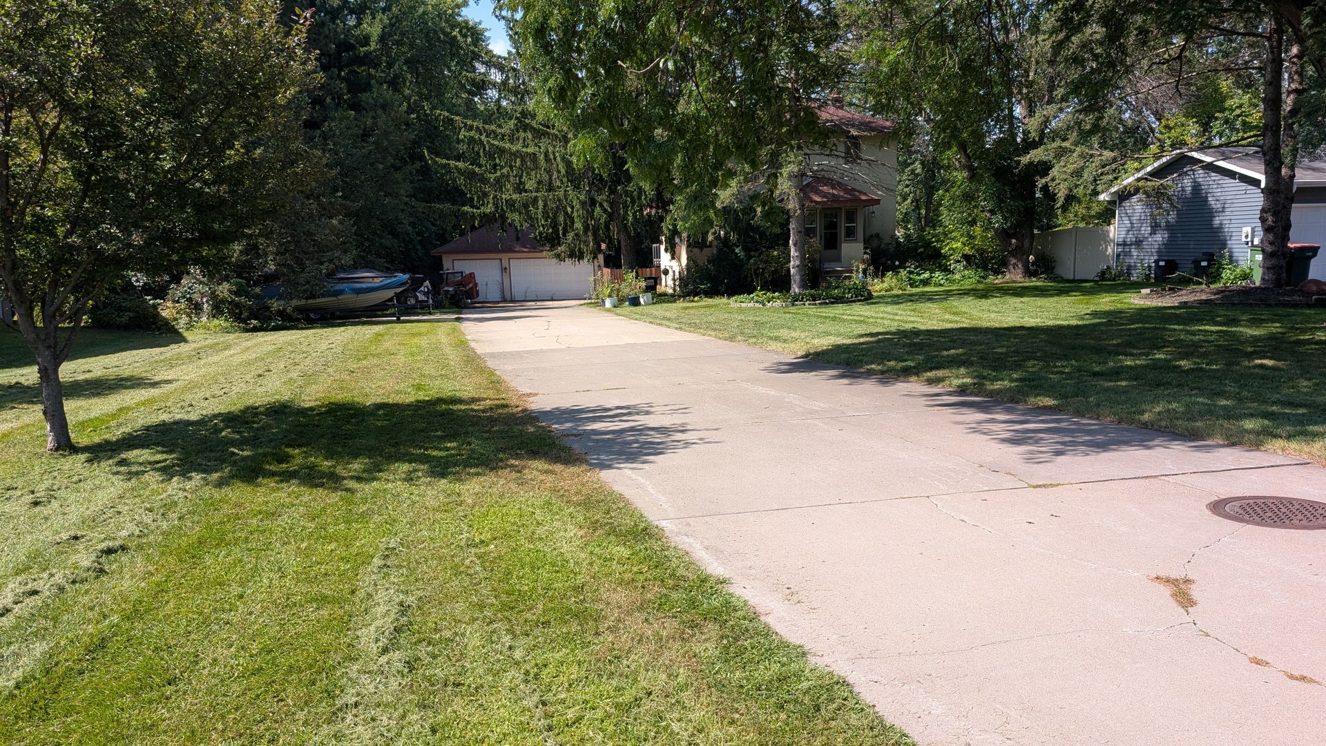 Driveway leading to a two-story house with a garage, trees, and a small shed on a grassy lawn.