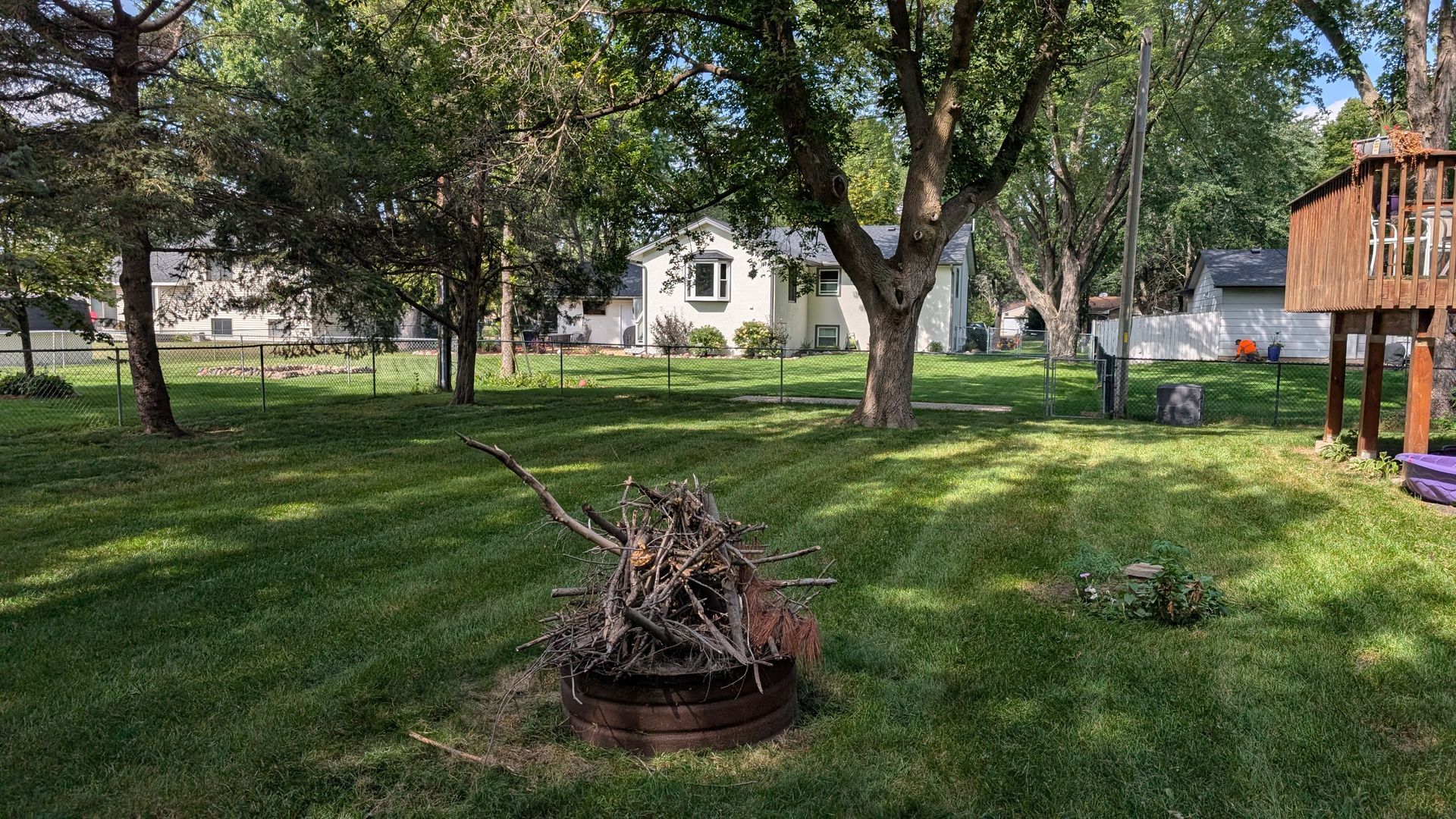 Backyard with trees, grass, a fire pit with wood, and a playhouse. A house is in the background.