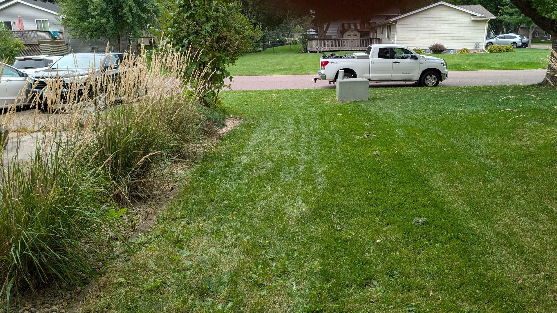 Green lawn with a white pickup truck driving on a residential street. Other vehicles parked along the curb.