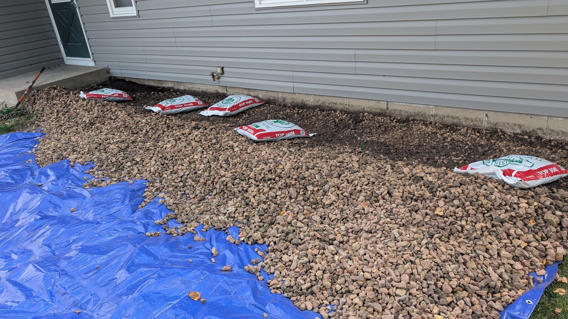 Brown mulch with bags sits along a house's foundation on a blue tarp.