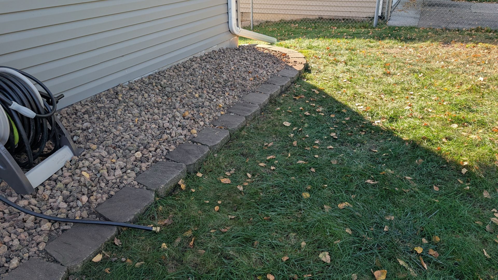 Gravel bed with stone border along the side of a house, hose reel, and grassy lawn.