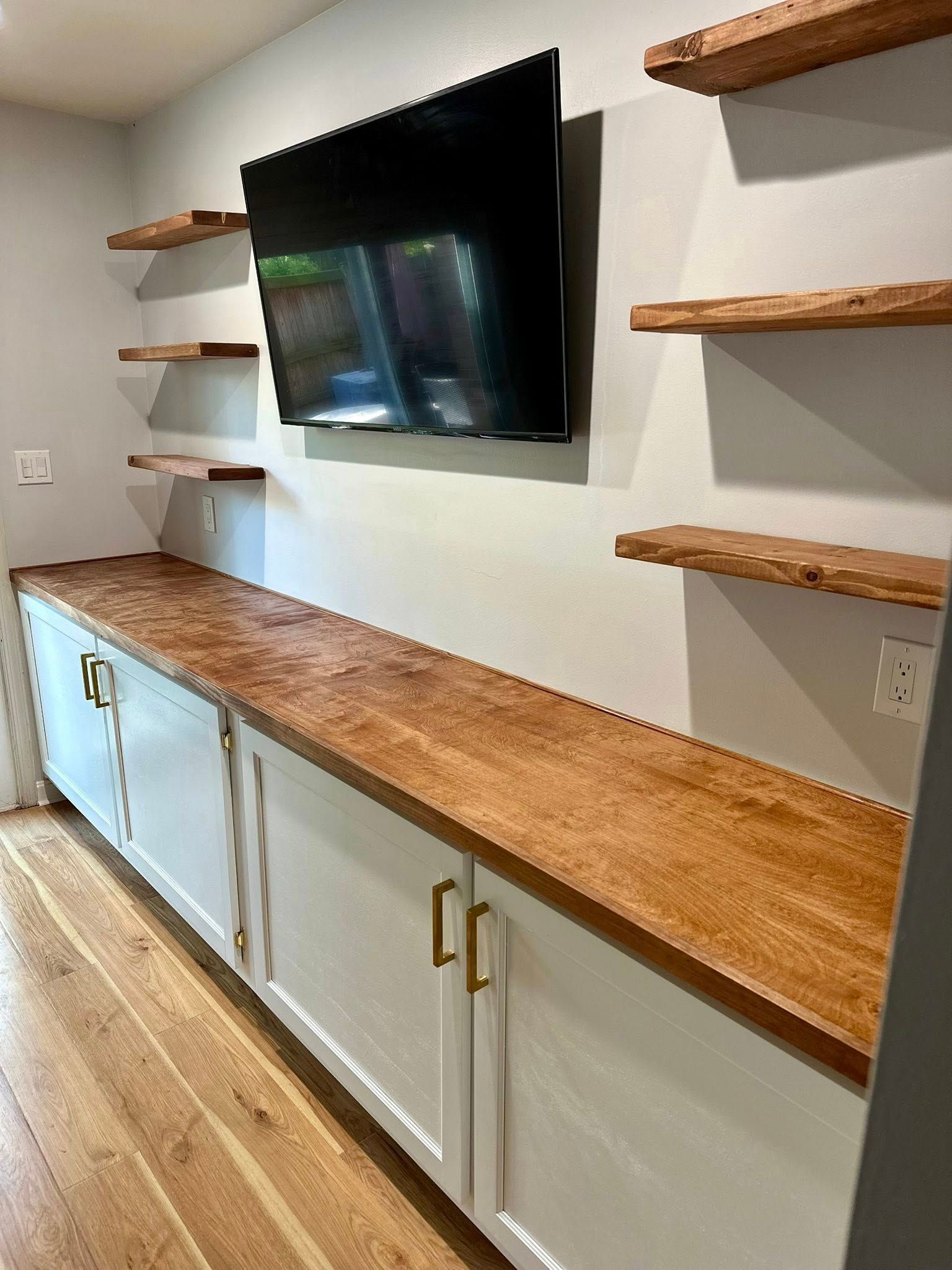 White cabinets with wooden countertop below a mounted TV and wooden shelves on a gray wall.