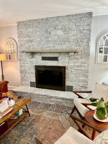 Stone fireplace with wooden mantel in a living room. Chairs and coffee table are in view.