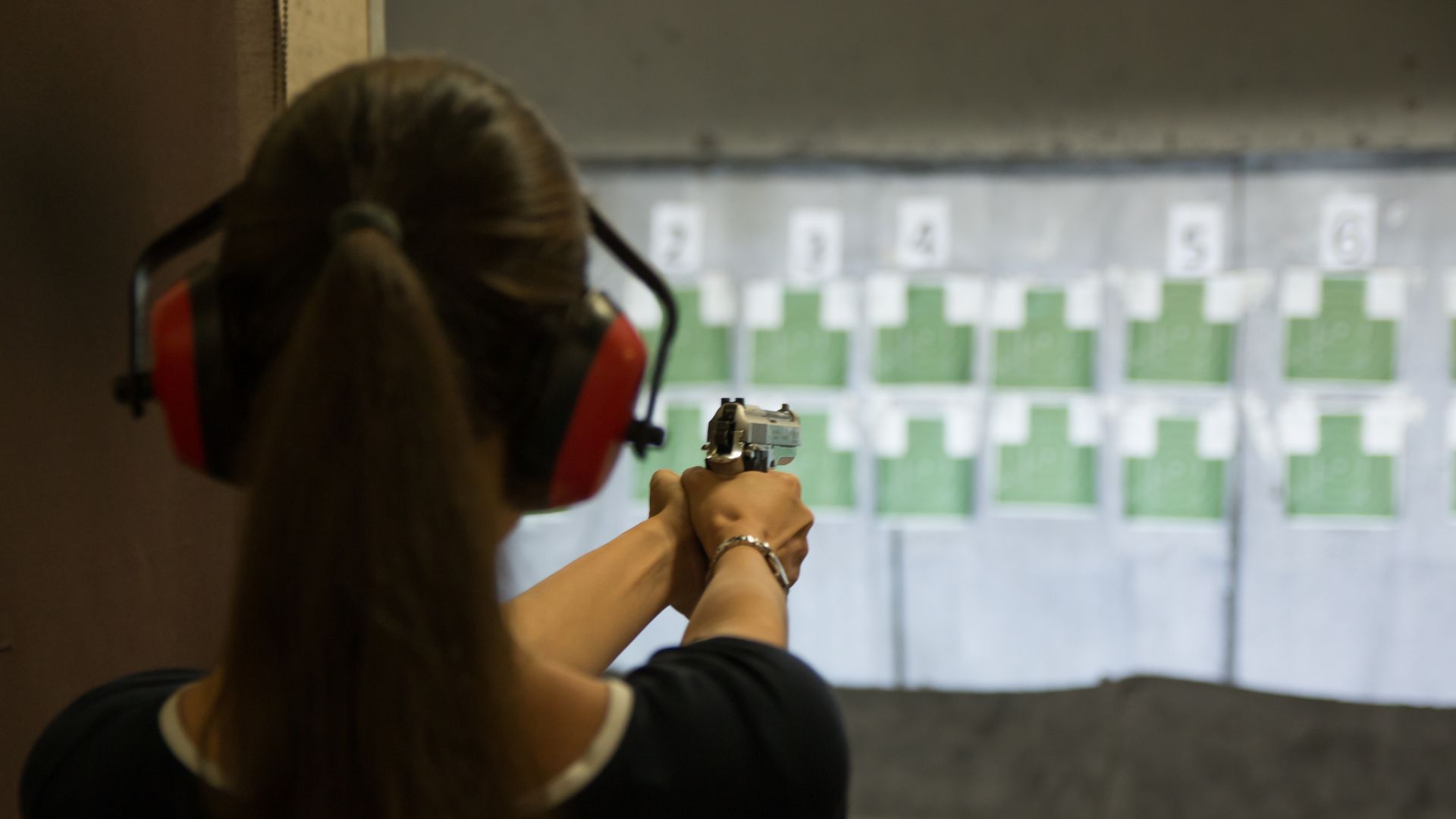 Person in a shooting range, aiming a handgun at a target. They wear ear protection, focusing intently.