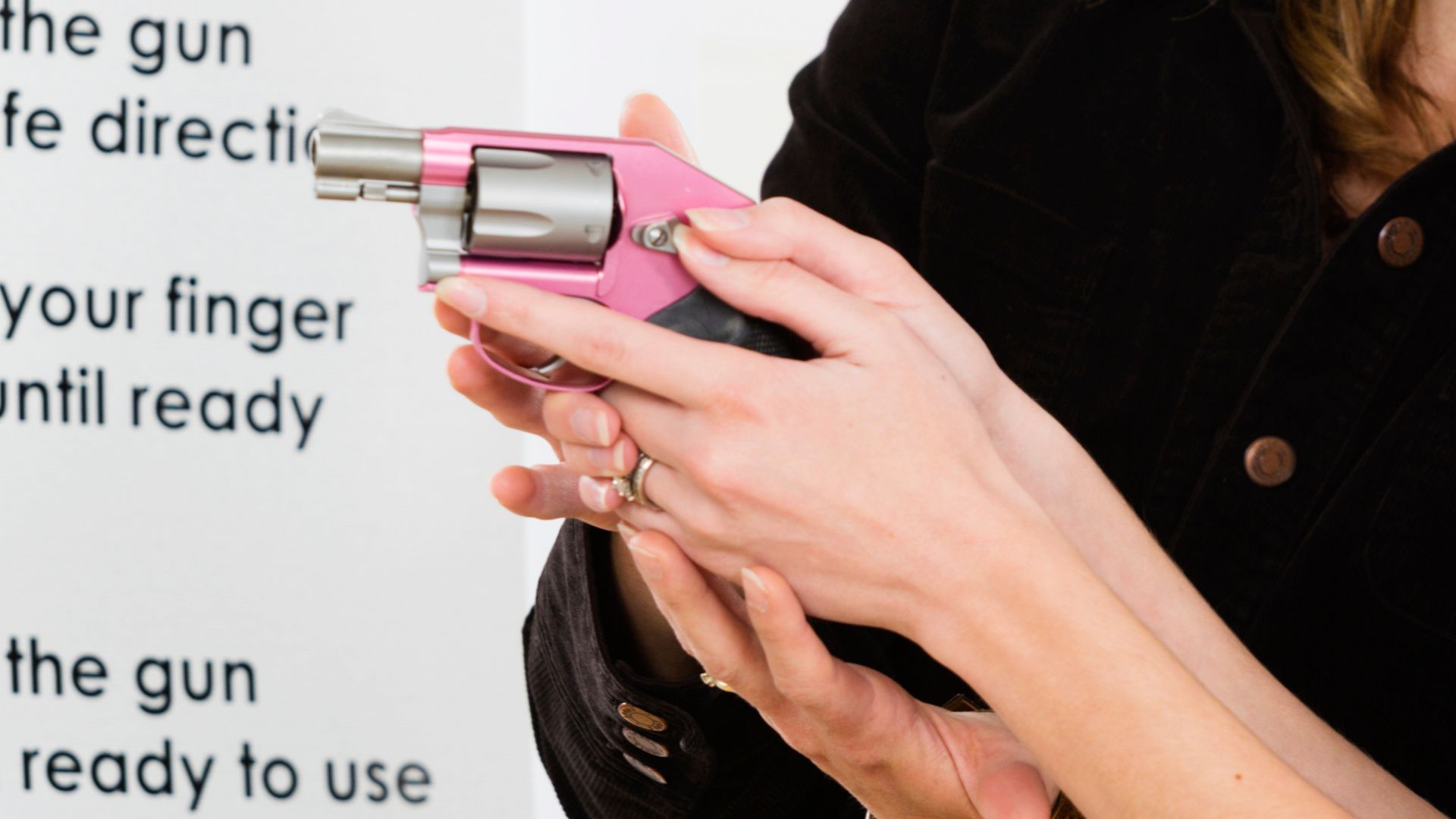 Person's hands holding a pink revolver, demonstrating safe gun handling near a safety poster.