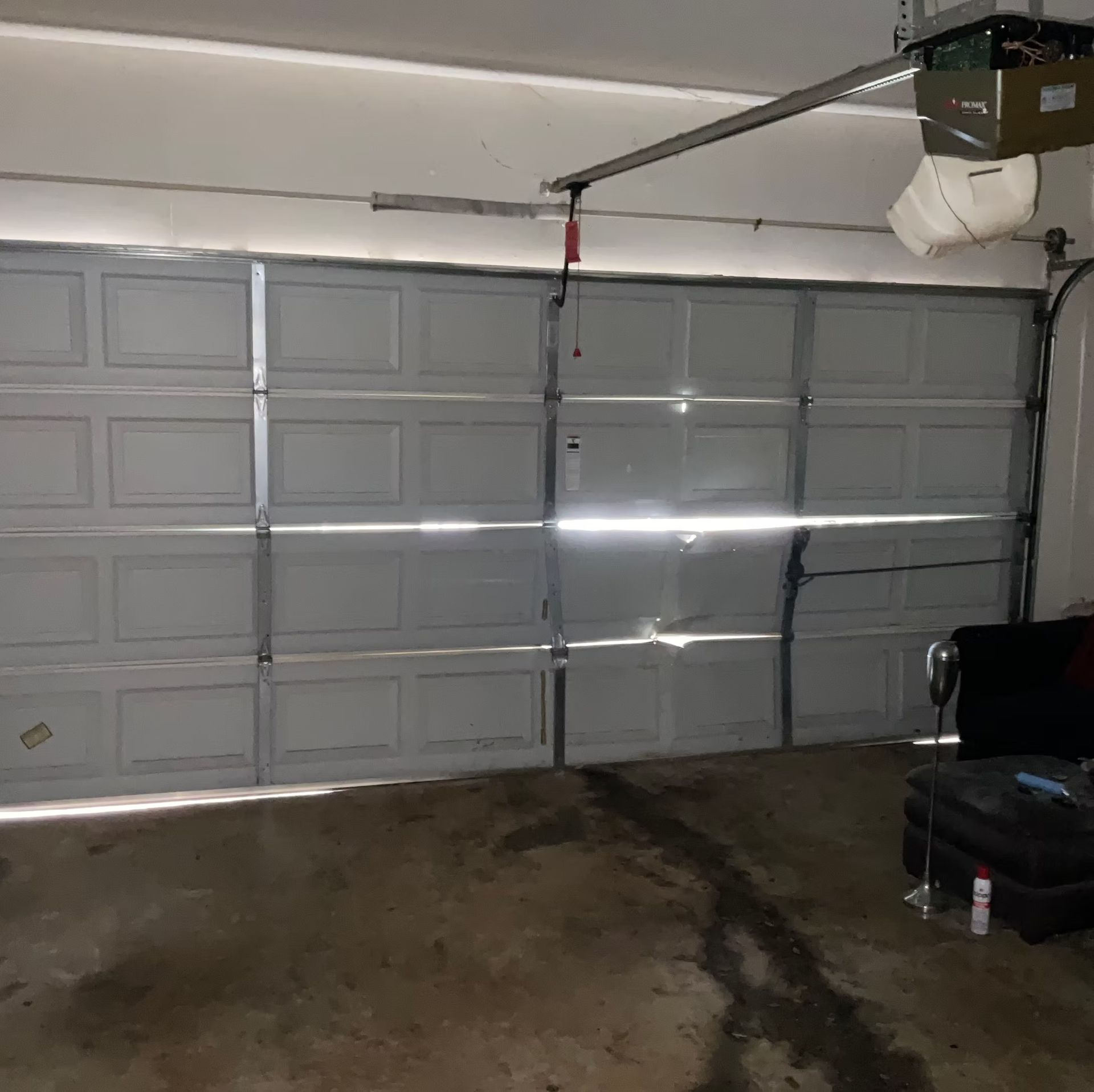 A partially open white garage door casting horizontal light, inside a dim garage with a stained concrete floor and a black couch.