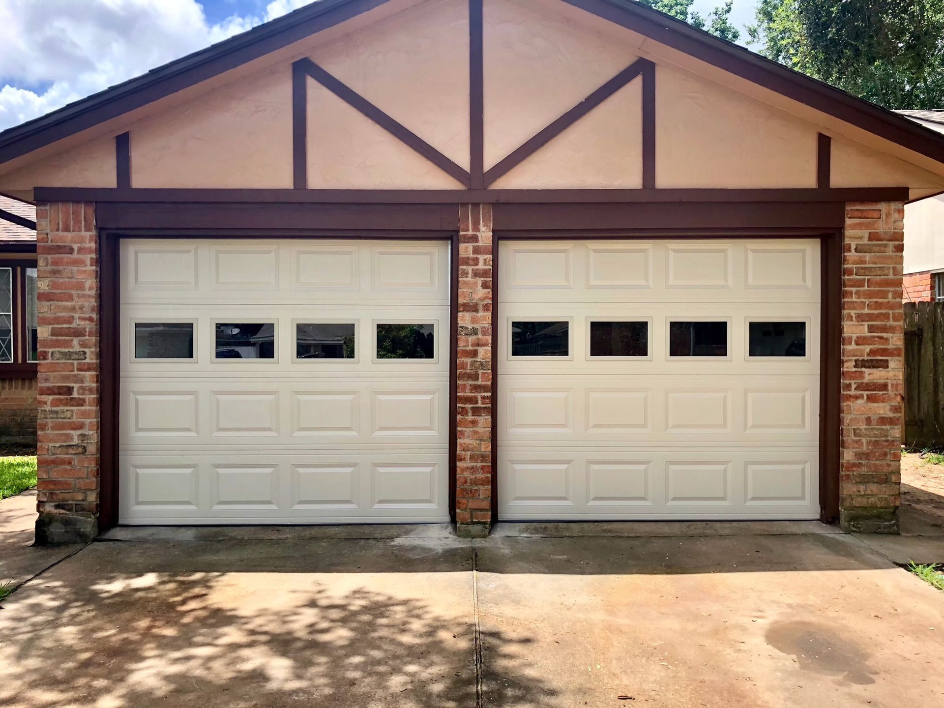 Two beige garage doors with small window panes, set in a brick and tan building. Brown trim accents the roof and doors.
