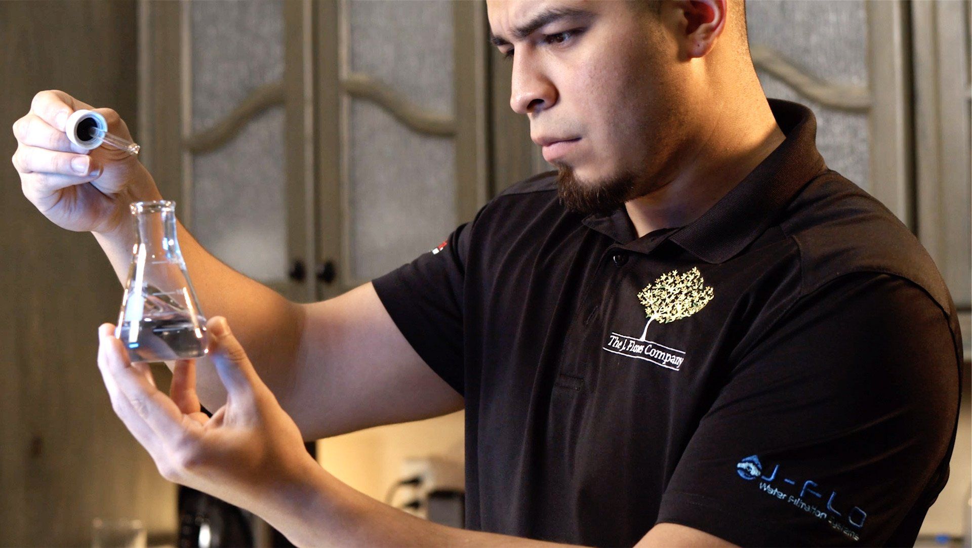 Man in black shirt with a goatee holding a flask, adding drops from a pipette. Indoors, cabinets in background.