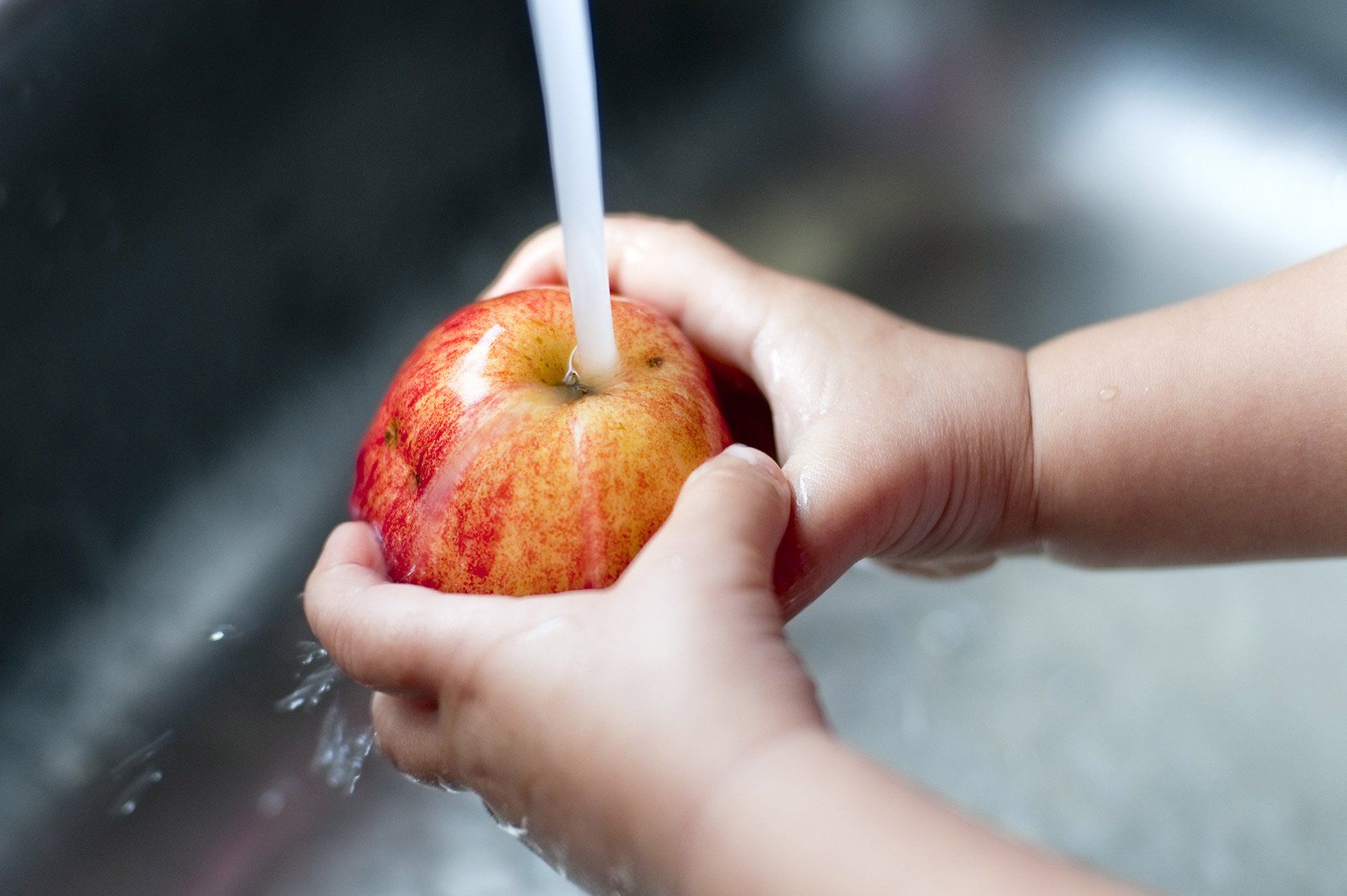Child washing a red and yellow apple under running water in a sink.