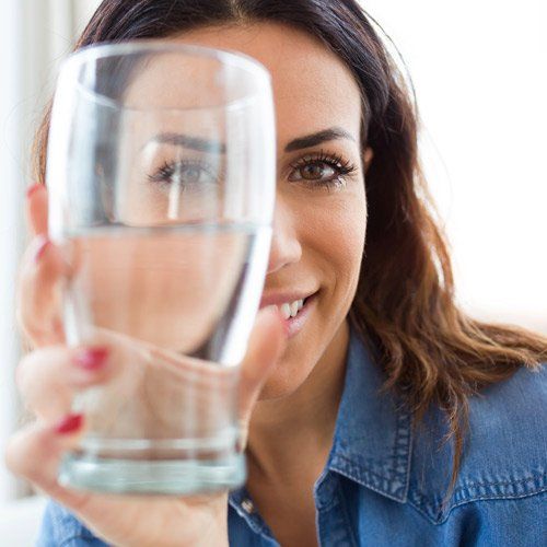 Woman smiling, holding a glass of water, looking at the camera. She has brown hair and is wearing a denim shirt.