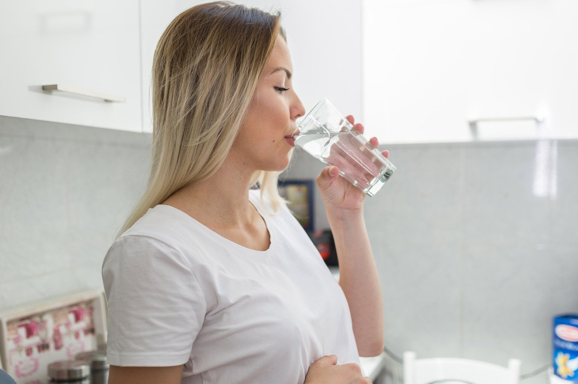 Woman in white shirt drinking water from a glass in a white kitchen.