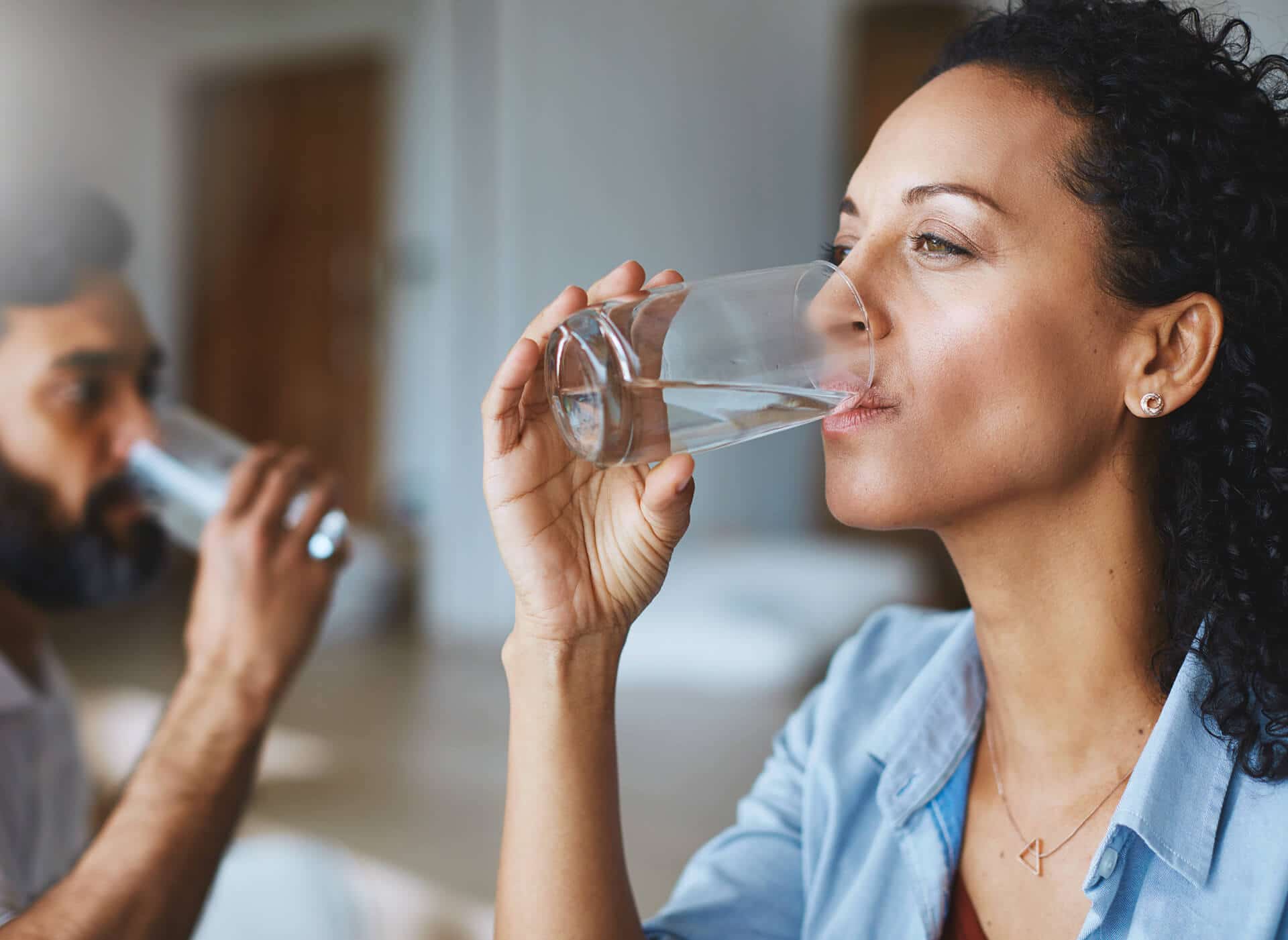 Woman drinks water from a glass; man in background also drinks. Indoors, sunny.