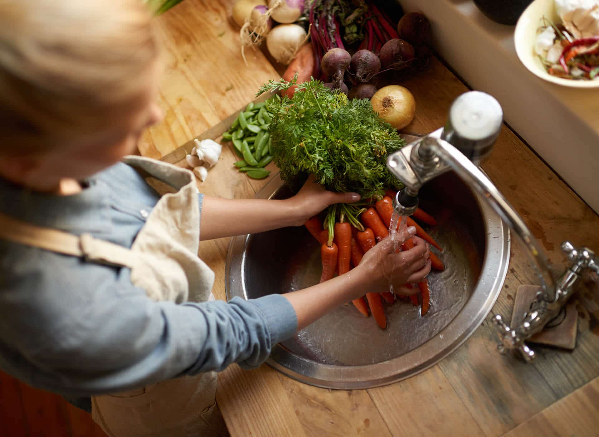 Woman washing carrots and herbs in a kitchen sink filled with water, other vegetables nearby.
