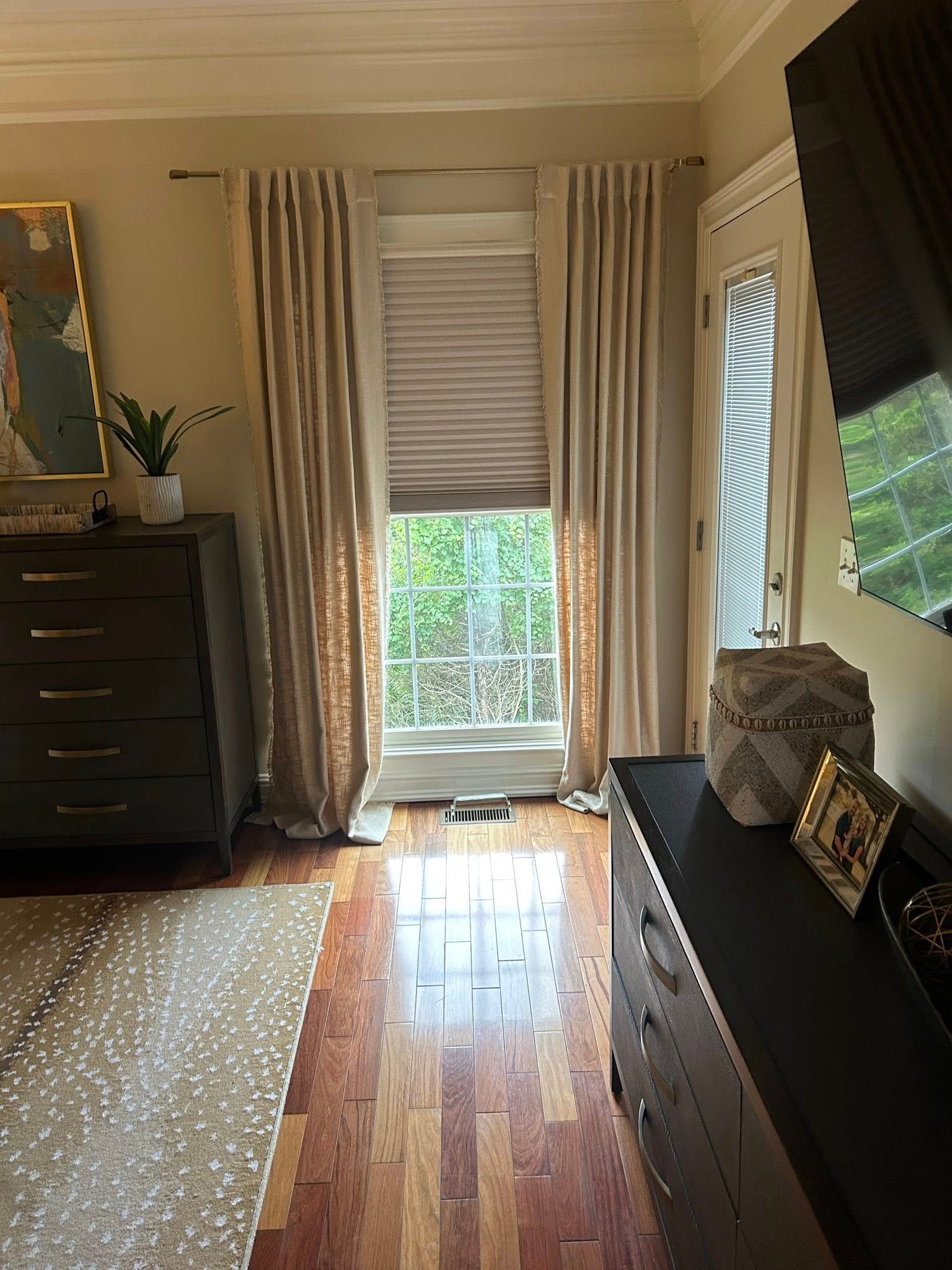 Bedroom interior with wood floors, tan curtains, and a window with blinds.