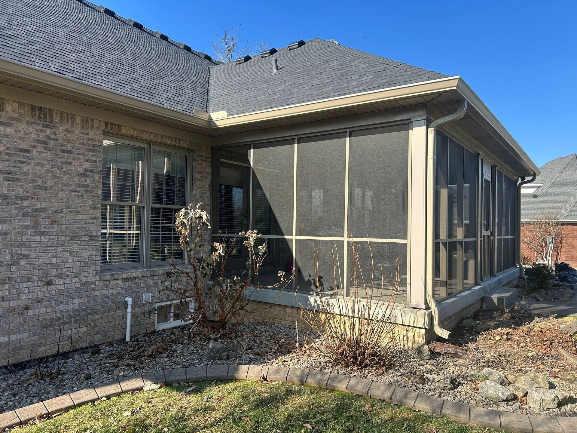 Screened porch attached to a brick house with a gray roof and gutter in a sunny outdoor setting.
