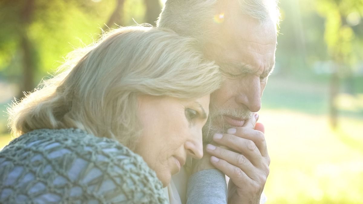 Two people resting close together outdoors in warm sunlight, one leaning on the other in a quiet embrace.