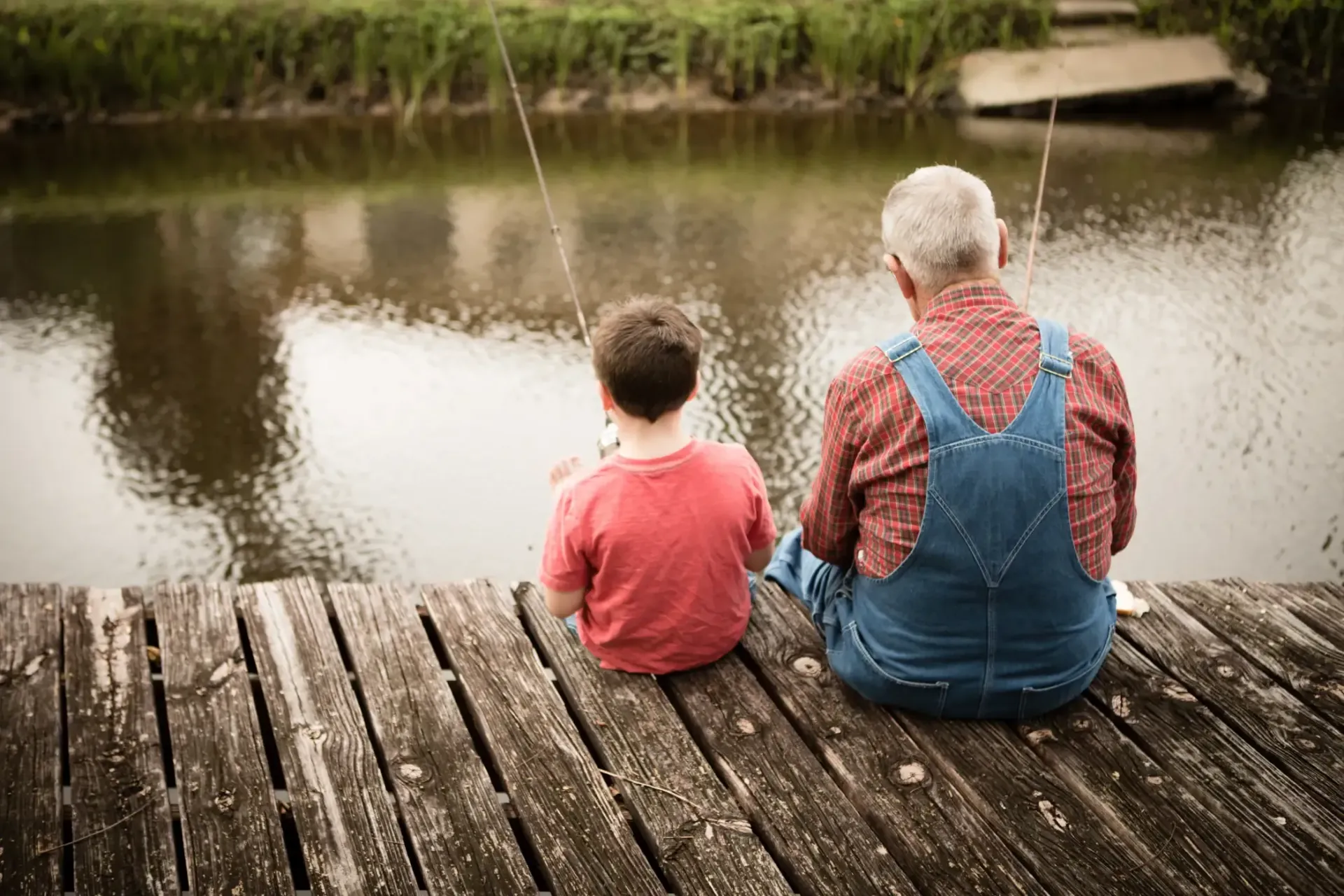Two people sit on a wooden dock, fishing beside a calm river.