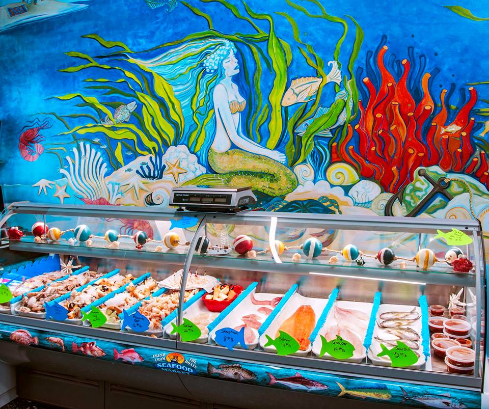 Seafood display case with fresh seafood, mermaid mural, and a scale in front of a blue wall.