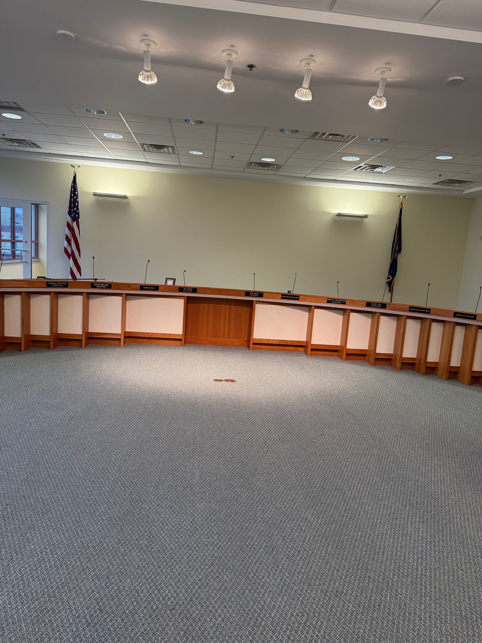 Empty meeting room with curved wooden table, two flags, and gray carpet.