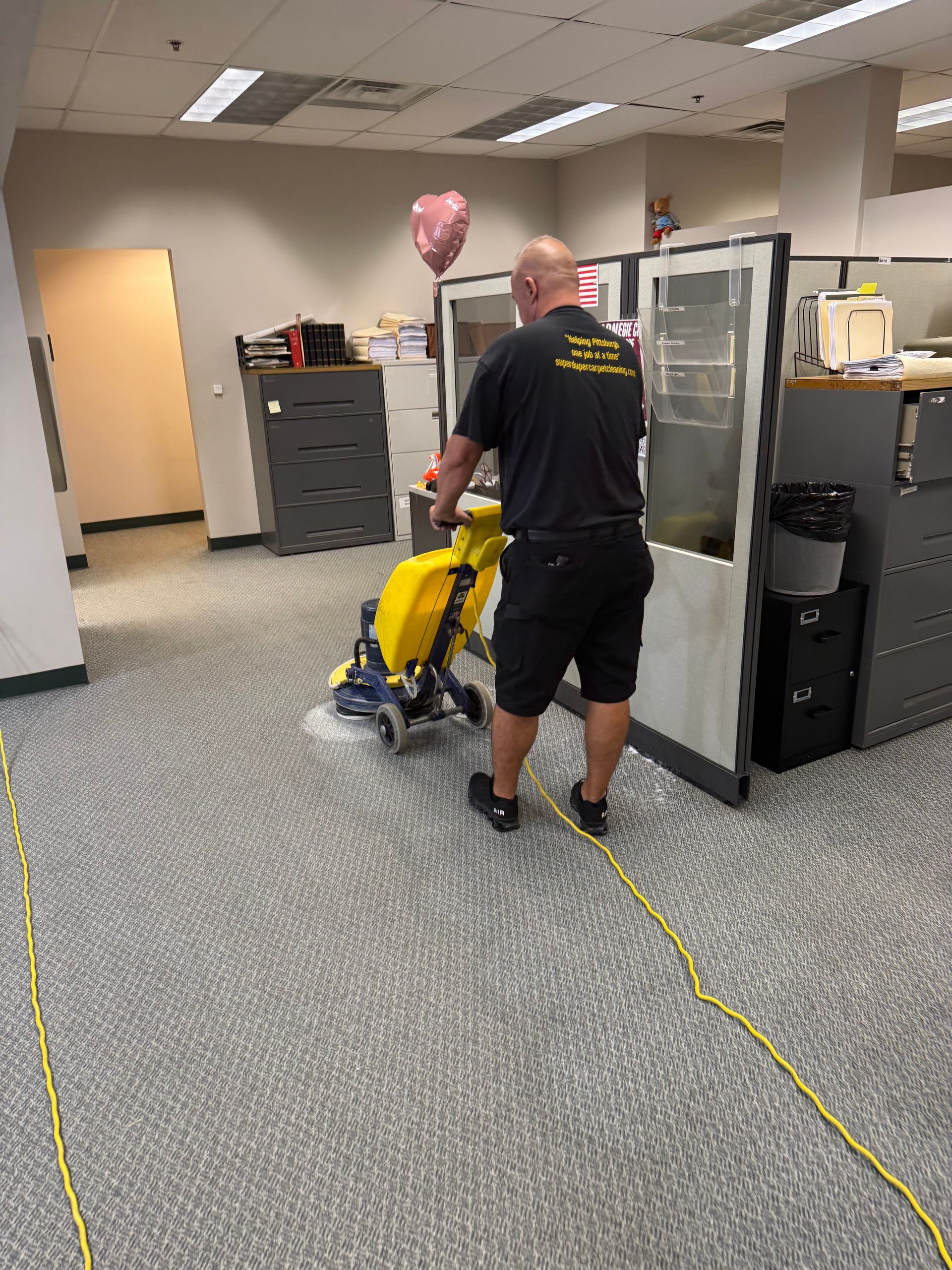 Man using a floor cleaning machine in an office with cubicles and gray carpet. Pink heart balloon is visible.
