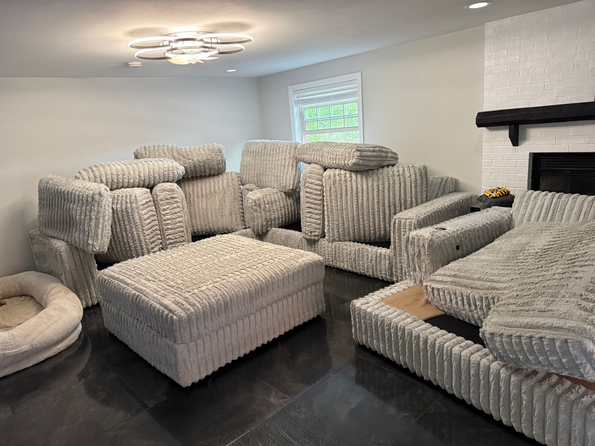 Living room with modular light gray sectional sofa. Ottoman, chair, and cushions are stacked. Dog bed in corner.