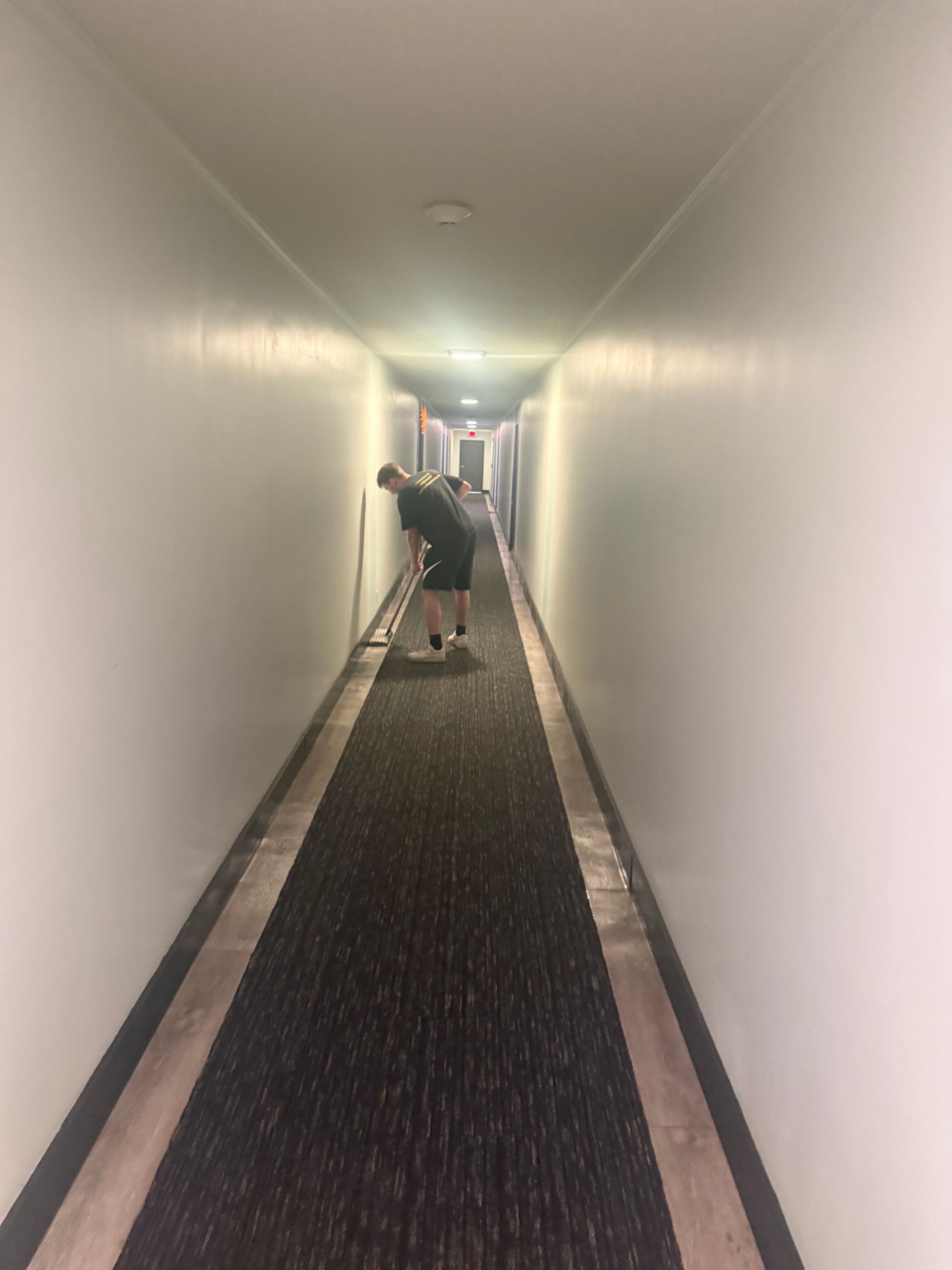 Person leaning in a long hotel hallway with dark carpet and white walls.