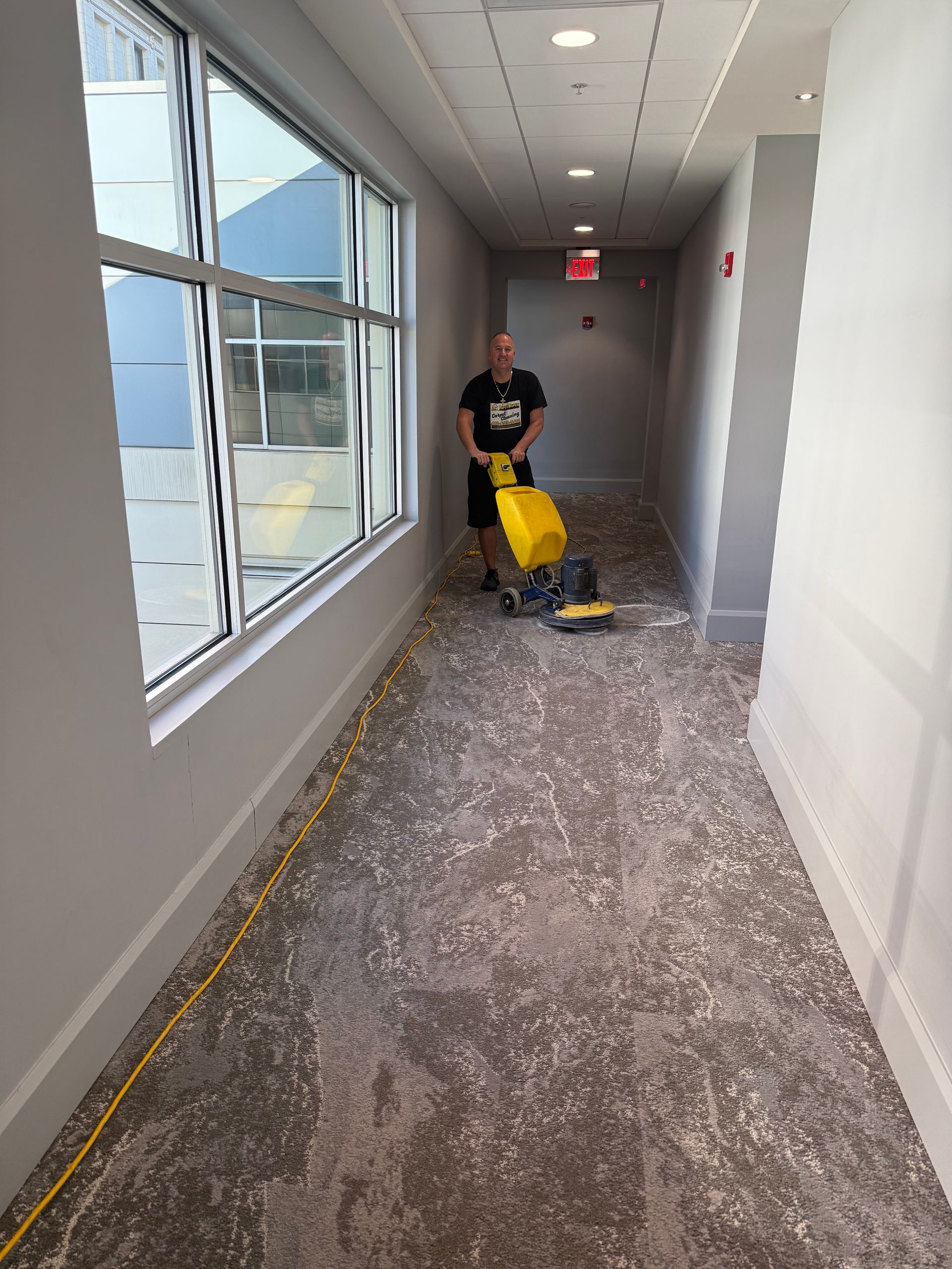 Person using a floor cleaning machine in a hallway with a large window. The floor is covered in patterned carpet.