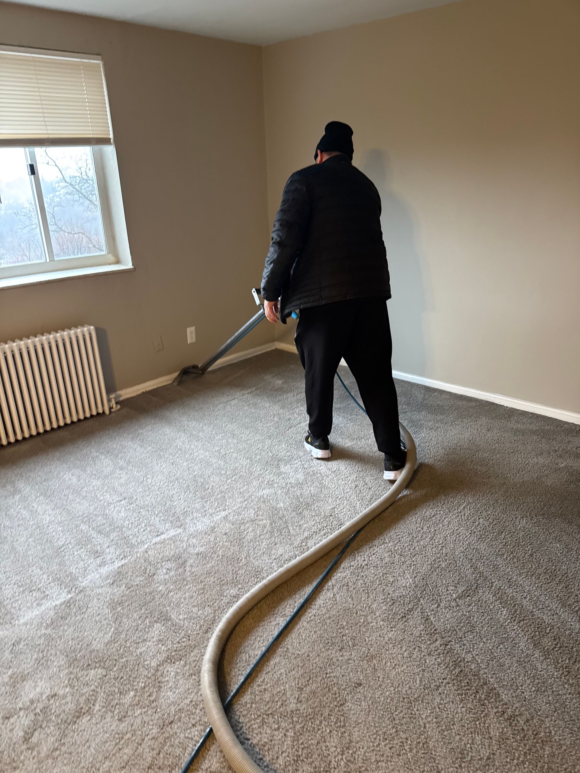 Person vacuuming gray carpet in a room with beige walls, a radiator, and a window.