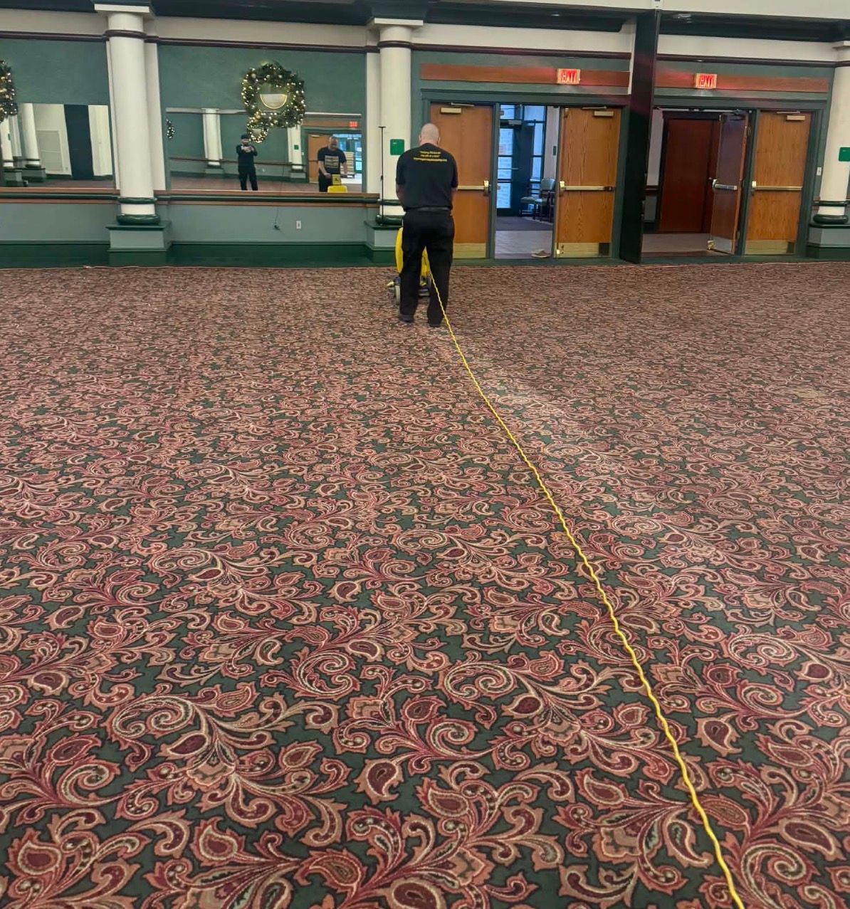 Person in a dark uniform using a measuring tape across a patterned carpeted floor in a large hall.