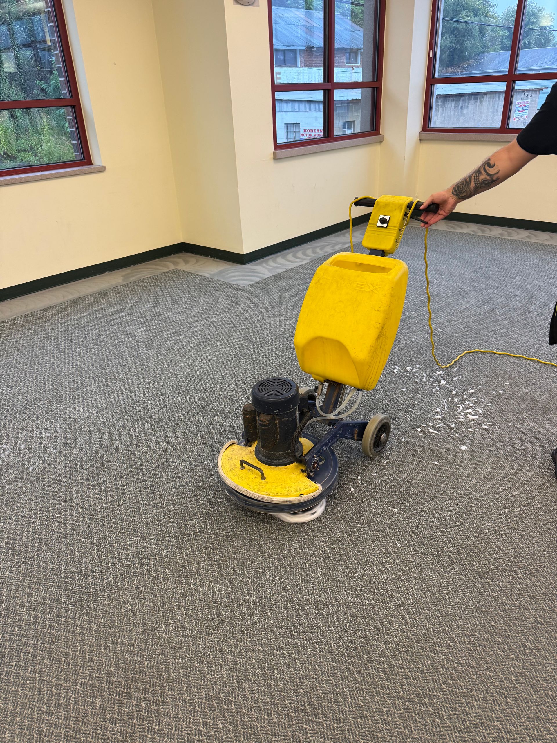 A person using a yellow carpet cleaning machine on a gray carpeted floor inside a room.