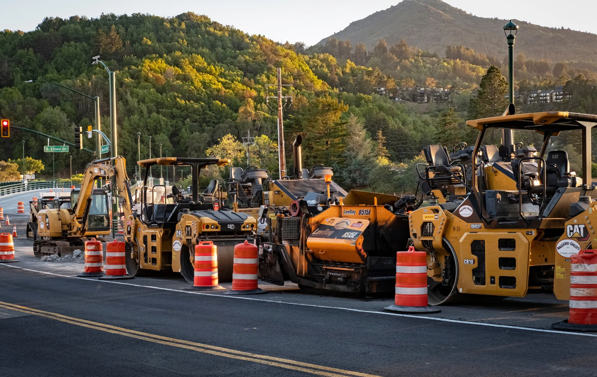 Construction equipment on a road, with orange barrels. Green trees and mountains in the background.