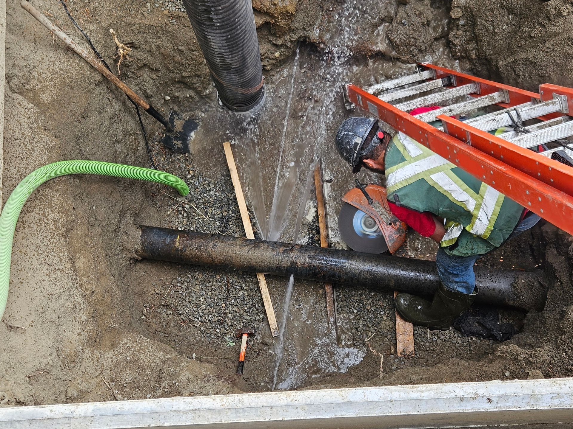 Construction worker cutting pipe in a trench, wearing safety vest and hard hat. Water sprays; orange ladder nearby.